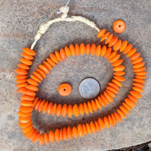 May include: A long strand of orange glass beads, strung together with a cream-coloured cord. The beads are shaped like teardrops and are arranged in a circular pattern. There are two smaller orange beads on the side.