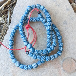 Puede incluir: Cadenas de cuentas a rayas azules y blancas, ensartadas en un cordón rojo. Las cuentas son redondas y tienen un diseño en espiral. Las cuentas son probablemente para la fabricación de joyas o manualidades. Cada cuenta mide aproximadamente 1 cm de diámetro.