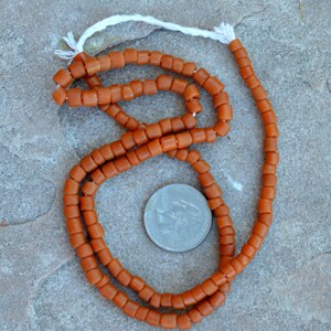 May include: A strand of orange-brown, barrel-shaped beads with a white string. A U.S. quarter is next to the beads for size comparison. The beads are arranged on a gray stone surface. The beads are likely for jewelry making.