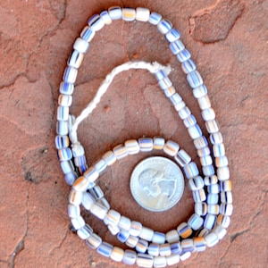 Puede incluir: Un collar hecho de cuentas de vidrio blancas, azules y amarillas. Las cuentas son pequeñas y de forma rectangular. El collar está enhebrado en un cordón blanco.