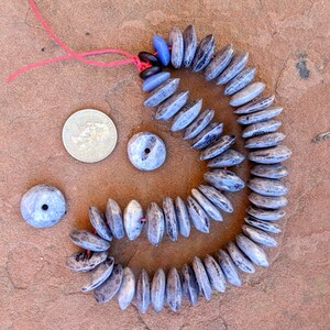May include: A strand of gray and blue gemstone beads with a red string threaded through the holes. The beads are oval shaped and have a rough texture. There are three loose beads next to the strand.