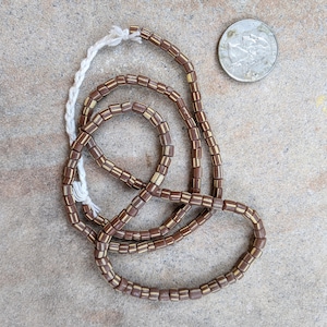 May include: A strand of brown and gold glass beads. The beads are square shaped and have a white stripe around the middle. The beads are strung on a white thread.