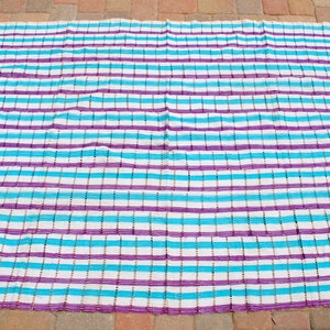 May include: A striped beach blanket with white, turquoise, and purple stripes. The blanket is made of a woven material and is perfect for relaxing on the beach or by the pool.