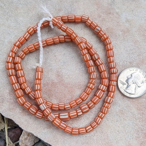 May include: A strand of orange and white striped glass beads. The beads are cylindrical and have a glossy finish. The beads are strung on a white cord.