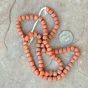 May include: A strand of orange and white striped glass beads. The beads are round and have a slightly irregular shape. The beads are strung on a white cord.