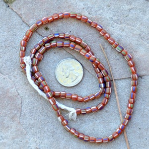 May include: A strand of orange, white, blue, and green glass beads. The beads are small and cylindrical. The strand is coiled around a US quarter.
