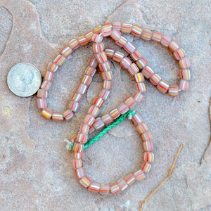 Könnte beinhalten: Eine Kette aus rosa, weißen und orangefarbenen Glasperlen mit einem grünen Faden, der durch sie gefädelt ist. Die Perlen sind quadratisch und haben ein gestreiftes Muster.