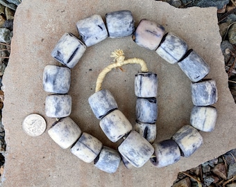 May include: A strand of white and grey bone beads, with a dark grey wash, arranged in a circle on a light brown surface. The beads are cylindrical and have a smooth, polished finish.