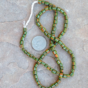 May include: A strand of green, orange, and white striped glass beads with a white string threaded through them. The beads are arranged in a circle and are lying on a brown, textured surface.