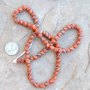May include: A strand of orange, white, and blue striped glass beads. The beads are round and have a slight swirl pattern. The beads are strung on a white thread.