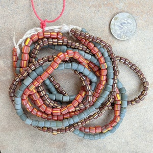 May include: A collection of vintage glass beads in shades of red, brown, and blue. The beads are strung together on a white cord and are arranged in a circular pattern.