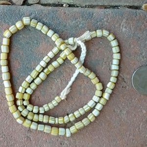 Puede incluir: Una hebra de cuentas de vidrio blancas y amarillas, enhebradas en un cordón blanco. Las cuentas son pequeñas y cilíndricas, con un patrón rayado.