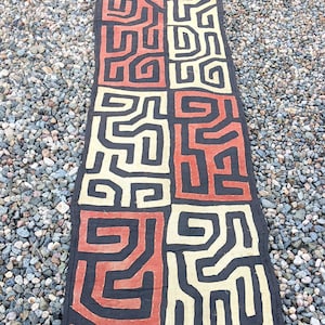 May include: A long, narrow, black fabric textile with a geometric pattern in red, orange, and white. The pattern is made up of repeating squares and rectangles.