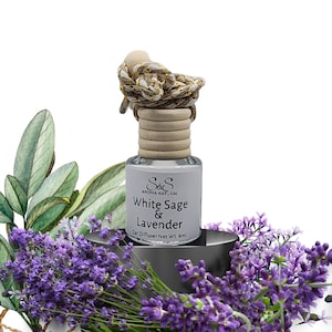 Può includere: Un diffusore per auto con un'etichetta bianca che recita "White Sage & Lavender". La bottiglia ha un tappo di legno e una corda decorativa. L'immagine è circondata da fiori di lavanda viola e foglie verdi.