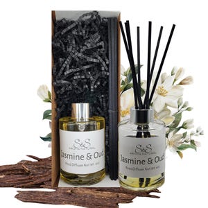 Może przedstawiać: Zestaw dyfuzora patyczkowego Jasmine & Oud. Zestaw zawiera dwie szklane butelki ze srebrnymi zakrętkami i białymi etykietami. W zestawie znajdują się również czarne patyczki i kwiaty dekoracyjne. Prezentowane w brązowym pudełku z czarnym papierem.
