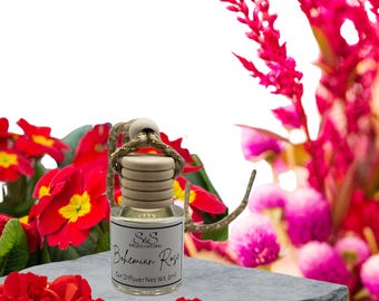 Puede incluir: Un pequeño ambientador para coche de cristal con tapón de madera y cuerda. La etiqueta dice "Bohemian Rose". El fondo presenta flores rojas y una superficie de piedra gris.