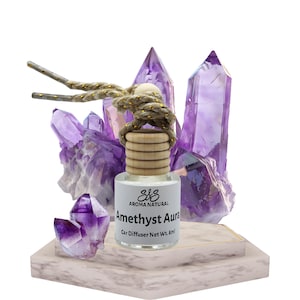 Op de afbeelding: Een auto luchtverfrisser met de tekst "Amethyst Aura" en een houten dop. Omringd door paarse amethist kristallen. Gepresenteerd op een marmer-patroon platform.