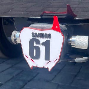 May include: A black trailer hitch with a white and red license plate-style tag. The tag has the name "SANDRO" above the number "61" in black. The hitch is attached to a dark-colored vehicle.
