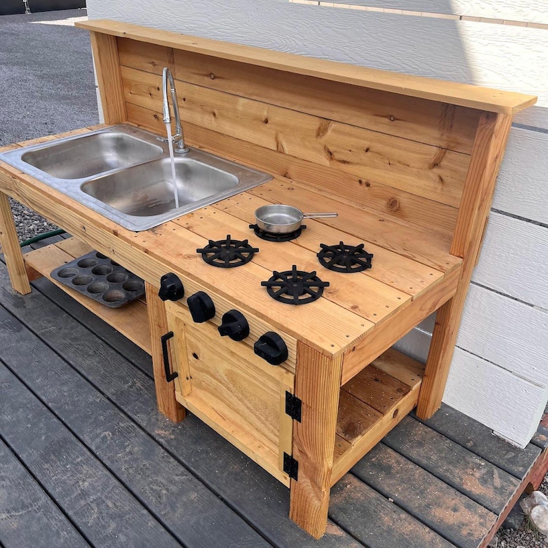 May include: A wooden play kitchen with a double sink, faucet, and stove top. The stove has four black burner grates and black knobs. A small metal pan and a muffin tin are also present. The kitchen is made of light-colored wood.