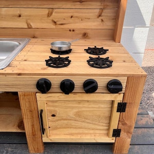 May include: A wooden play kitchen with a stainless steel sink, four black burner grates, and four black knobs. A small metal pan sits on one of the burners. The kitchen has a cabinet with a black handle and hinges.