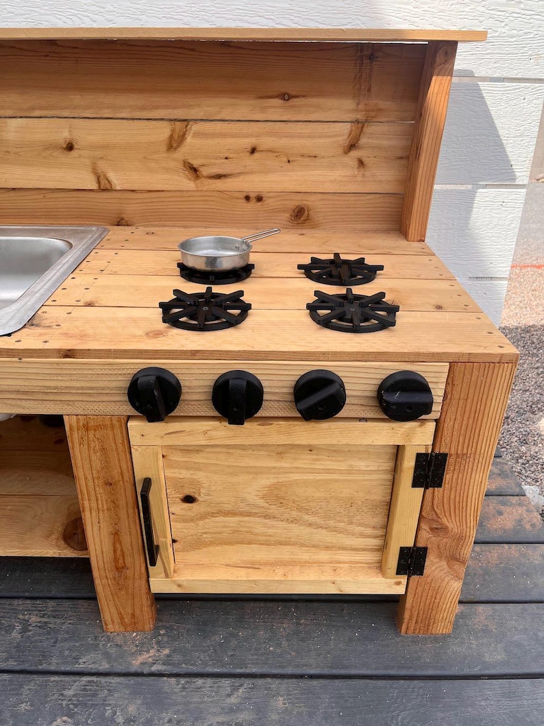 May include: A wooden play kitchen with a stainless steel sink, four black burner grates, and four black knobs. A small metal pan sits on one of the burners. The kitchen has a cabinet with a black handle and hinges.