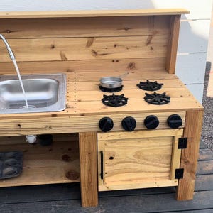May include: A wooden outdoor play kitchen with a double sink, faucet, and stove top. The stove has four black burner grates and four black knobs. A small metal pan sits on the stove. The kitchen is made of light-colored wood.