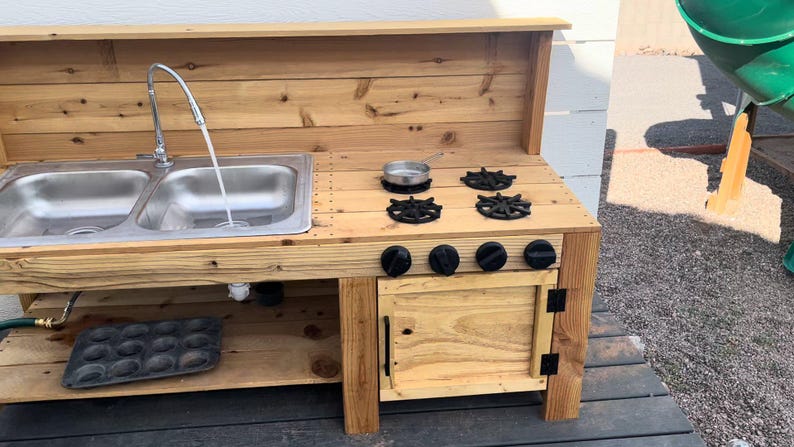 May include: A wooden outdoor play kitchen with a double sink, faucet, and stove top. The stove has four black burner grates and four black knobs. A small metal pan sits on the stove. The kitchen is made of light-colored wood.