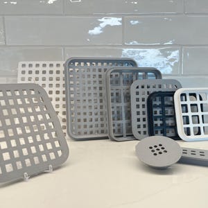 Puede incluir: Un juego de nueve escurridores de platos de plástico gris y blanco. Los escurridores son cuadrados y tienen un patrón de rejilla. También hay un tapón de drenaje de plástico gris con un patrón de rejilla.