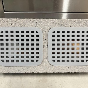May include: Two gray, square, slotted vent covers are set into a gray cinder block. The vent covers have rounded corners and a grid pattern of small square openings. The cinder block has a rough, textured surface.
