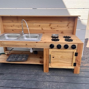 May include: A wooden outdoor play kitchen with a double sink, faucet, and stovetop. The kitchen features a small cabinet with a door and black knobs. A small pot sits on the stovetop. The kitchen is made of light-colored wood.