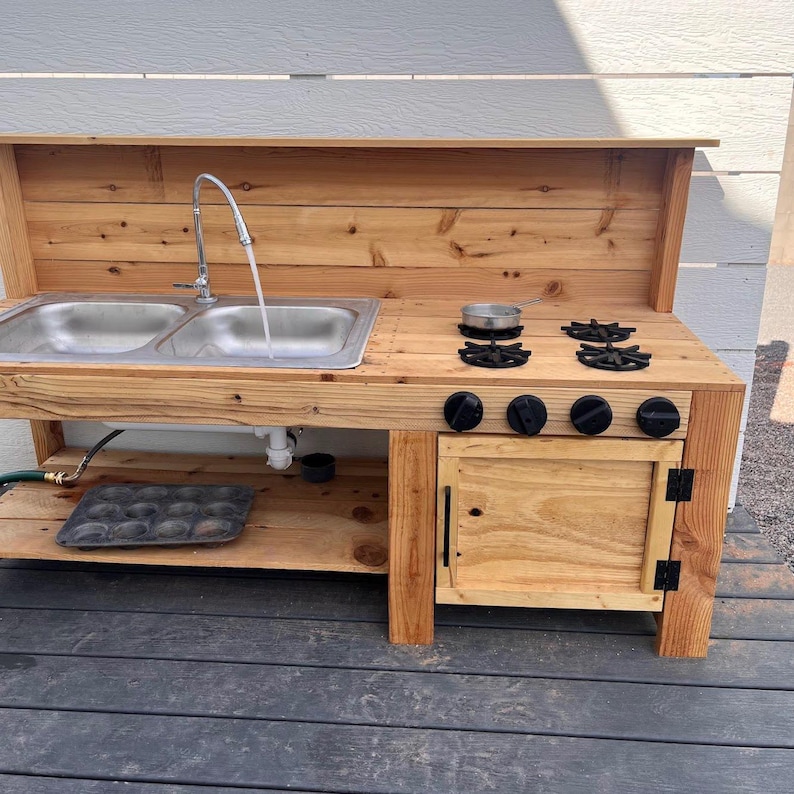 May include: A wooden outdoor play kitchen with a double sink, faucet, and stovetop. The kitchen features a small cabinet with a door and black knobs. A small pot sits on the stovetop. The kitchen is made of light-colored wood.