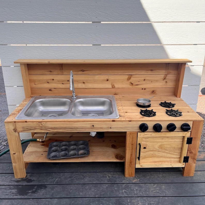 May include: A wooden outdoor play kitchen with a double sink, faucet, and stove top. The kitchen features a small oven, black knobs, and a shelf for storage. The natural wood finish gives it a rustic look.