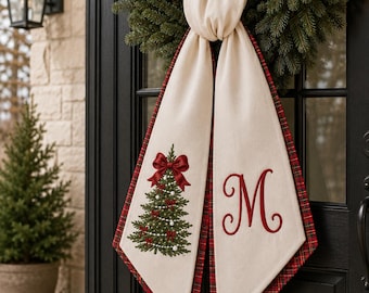 May include: A cream-colored fabric door hanger featuring a red and green plaid backing. One side displays an embroidered green Christmas tree with a red bow and white ornaments, while the other side features a large, embroidered red letter M.