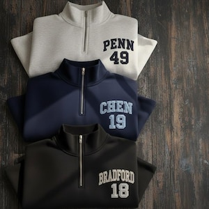 May include: Three quarter-zip pullovers in grey, navy, and black are neatly arranged. Each features a contrasting embroidered name and number: "PENN 49", "CHEN 19", and "BRADFORD 18". The pullovers are made of a soft material.