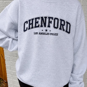 May include: A light gray crewneck sweatshirt featuring the dark navy blue text CHENFORD in an arched collegiate font, followed by three small stars and the text LOS ANGELES POLICE in a smaller, straight, dark navy blue sans-serif font.