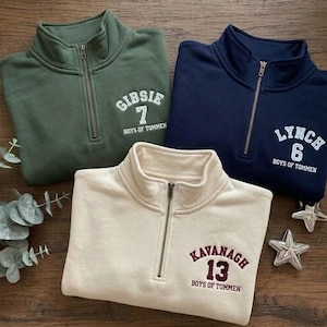 May include: Three quarter-zip sweatshirts in olive green, navy blue, and cream. Each sweatshirt features a name and number, with the text "BOYS OF TOMMEN" below. The sweatshirts are displayed on a wooden surface.