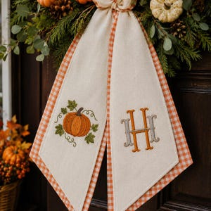May include: A decorative fabric door hanger featuring two cream-coloured panels with orange and white gingham trim. One panel displays an embroidered orange pumpkin with green leaves, while the other features an embroidered monogram letter H.