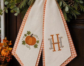 May include: A decorative fabric door hanger featuring two cream-colored panels with orange and white gingham trim. One panel displays an embroidered orange pumpkin with green leaves, while the other features an embroidered monogram letter H.