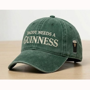 Könnte beinhalten: Eine grüne Baseballkappe mit dem gestickten Text "DADDY NEEDS A GUINNESS" in Weiß. Auf der Seite befindet sich ein kleines gesticktes Bild eines Guinness-Pints. Die Kappe hat einen gebogenen Schirm und einen Knopf oben.