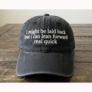 Puede incluir: Gorra de béisbol gris oscuro con texto bordado en blanco: "i might be laid back but i can lean forward real quick". La gorra tiene una visera curva y un botón en la parte superior.