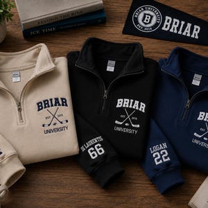 May include: Three quarter-zip sweatshirts in beige, black, and navy blue, each embroidered with "Briar University" and crossed hockey sticks. The beige sweatshirt has "Graham 44" on the sleeve, black has "Di Laurentis 66", and navy has "Logan 22". A Briar pennant is also visible.