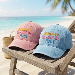 May include: A pink and a light blue baseball cap, each featuring the embroidered text Summer Should Be Fun! in a multi-coloured font with a small white flower icon replacing the letter O. Both caps have a curved brim and a structured crown.