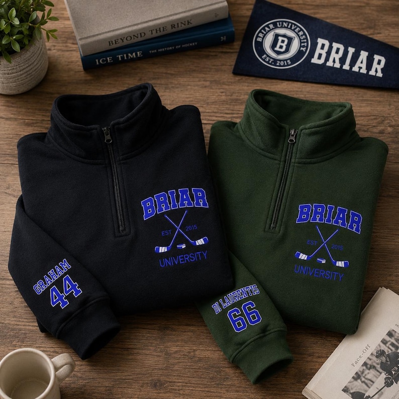 May include: Two quarter-zip pullovers, one black and one green, with embroidered "Briar University" logos and hockey stick graphics. The black pullover has "Graham 44" on the sleeve, and the green one has "St Laurentis 66".