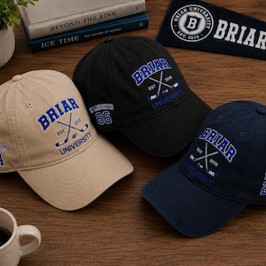 May include: Three baseball caps in tan, black, and navy blue, each embroidered with "Briar University" and crossed hockey sticks. The tan cap has "Graham 44" on the side, the black cap has "66", and the navy cap has "Logan 22".