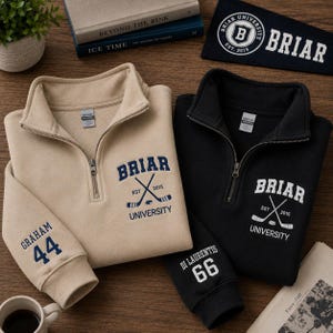 May include: Two quarter-zip sweatshirts, one tan and one black, with the text "BRIAR UNIVERSITY" and crossed hockey sticks. The tan sweatshirt has "GRAHAM 44" on the sleeve, while the black one has "DI LAURENTIS 66". A pennant and books are in the background.