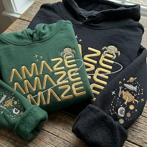 Könnte beinhalten: Zwei Kapuzenpullover, einer dunkelgrün und einer schwarz, mit goldgesticktem Text und Weltraumgrafiken. Der grüne Pullover sagt "AMAZE" und der schwarze "ZEN". Beide haben Schildkröten- und Sternbilddesigns.