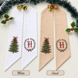 May include: Two pairs of long fabric ribbons in white and sand colors. Each ribbon features an embroidered green Christmas tree with a red bow or a red letter H inside a green wreath. Gold-colored metal bells are attached to the top of each pair.