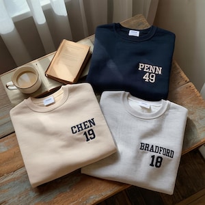 May include: Three folded crewneck sweatshirts in beige, navy blue, and light gray. Each sweatshirt has a different name and number embroidered on the front: "CHEN 19", "PENN 49", and "BRADFORD 18". A book and a mug of coffee are in the background.