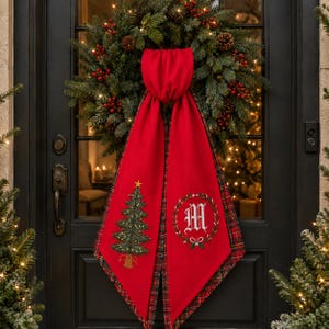 May include: A red fabric door hanger featuring a green embroidered Christmas tree on one side and a white letter M inside a green wreath on the other. The hanger has a red and green plaid lining and is draped over a green pine wreath with red berries.
