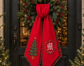 May include: A red fabric door hanger featuring a green embroidered Christmas tree on one side and a white letter M inside a green wreath on the other. The hanger has a red and green plaid lining and is draped over a green pine wreath with red berries.